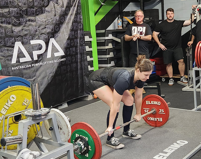 Lady Performing a Deadlift at a Darwin Powerlifting Competition with Sponsors in the background.